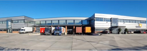 Bedrijfspand van exotisch voedselspecialist Unidex. Je ziet een groot zilvergrijs langgerekt gebouw met aan de uiteinden de kantoorpanden en in het midden het laadplatform waar enkele vrachtwagens aan het lossen en laden zijn.