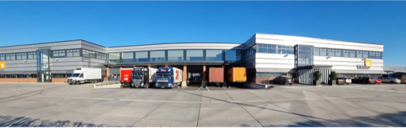 Bedrijfspand van exotisch voedselspecialist Unidex. Je ziet een groot zilvergrijs langgerekt gebouw met aan de uiteinden de kantoorpanden en in het midden het laadplatform waar enkele vrachtwagens aan het lossen en laden zijn.