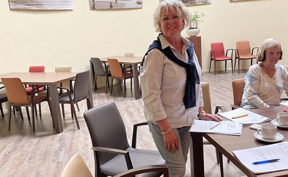 Vrouw met grijs golvend haar tot net na de oren, pony. Ze draagt een witte blouse, lichte spijkerbroek en om haar schouders een donkerblauwe trui geknoopt. Ze kijkt vriendelijk lachend de camera in.