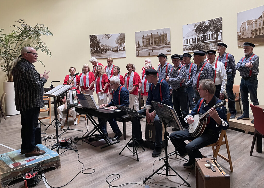 Foto van een optreden van De Ringvaartzangers na de ALV van VVS Hillegom april 2026. Rechts mannen in een visserskostuum met donkere broek en pet en blauw gestreept overhemd en rode bretels. Links mannen en vrouwen in een licht beige broek, rood jasje en lichtblauw sjaaltje.