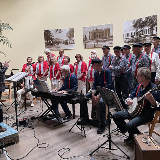 Foto van een optreden van De Ringvaartzangers na de ALV van VVS Hillegom april 2026. Rechts mannen in een visserskostuum met donkere broek en pet en blauw gestreept overhemd en rode bretels. Links mannen en vrouwen in een licht beige broek, rood jasje en lichtblauw sjaaltje.