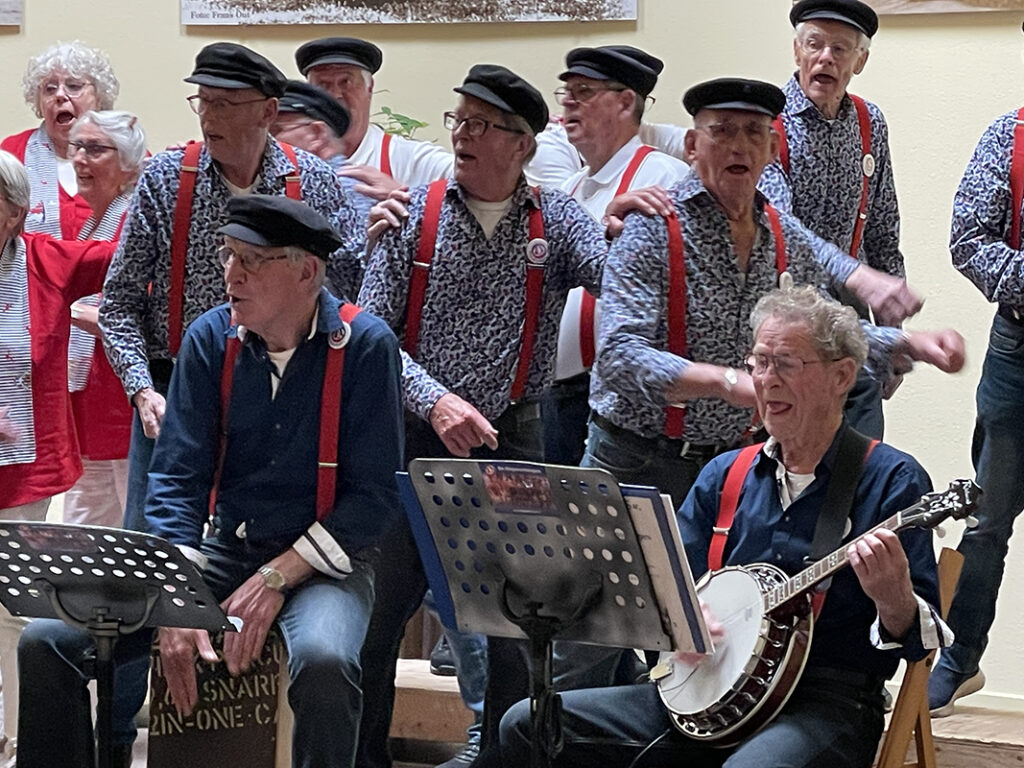 Close-up van enthousiaste koorleden van de Ringvaartzangers tijdens optreden bij VVS Hillegom. Mannen dragen visserspetten en blauw bloesje met rode brettels. Mannen hebben hand op schouder persoon voor hun. Voor de zangers een muzikant met banjo.