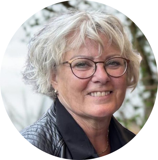 Vrouw van seniore leeftijd met grijs golvend haar tot onderaan oren met pony. Ze draagt een fijne zwarte ronde bril, een zwart leren jack en zwarte blouse.