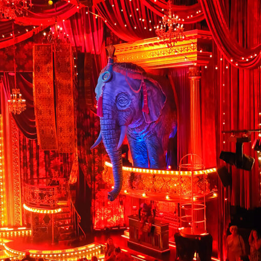 Decor musical Moulin Rouge in Beatrix Theater. Je ziet een grote geprojecteerde olifant op een balkon in rood verlichte zaal.
