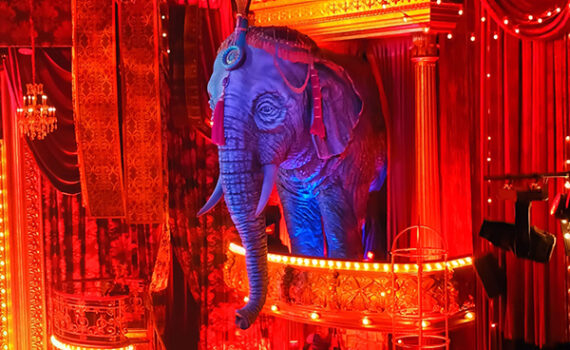 Decor musical Moulin Rouge in Beatrix Theater. Je ziet een grote geprojecteerde olifant op een balkon in rood verlichte zaal.